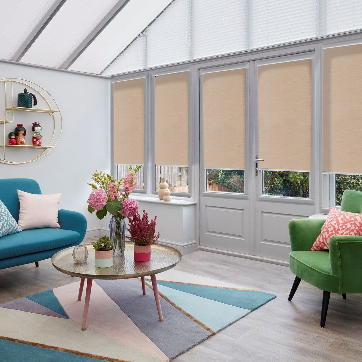 Beige roller blinds across tall conservatory windows and French doors, partially lowered and filtering daylight; bright sunroom with teal sofa, green armchair, coffee table, pastel geometric rug, and garden glimpses.