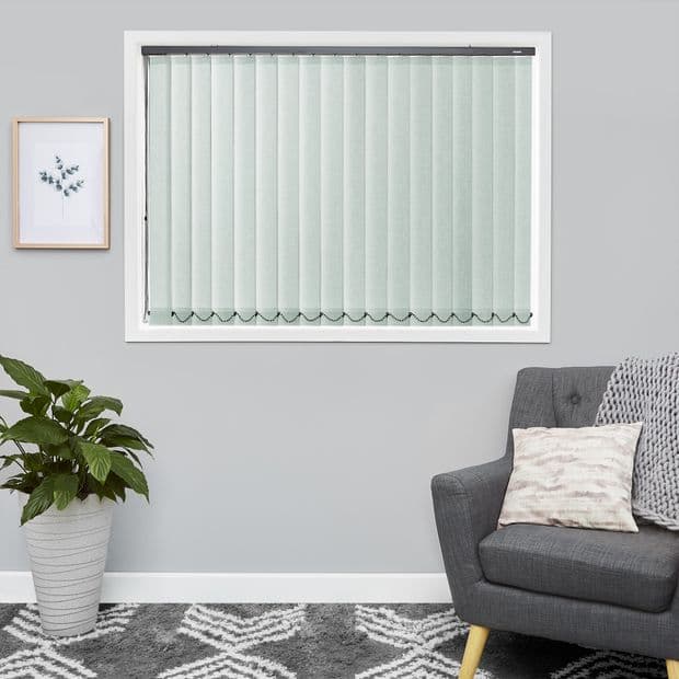 Vertical fabric blinds, pale mint, full-length slatted panels with scalloped weighted chains at bottoms, lowered and closed, filtering soft daylight in a contemporary living room with gray armchair and plant.