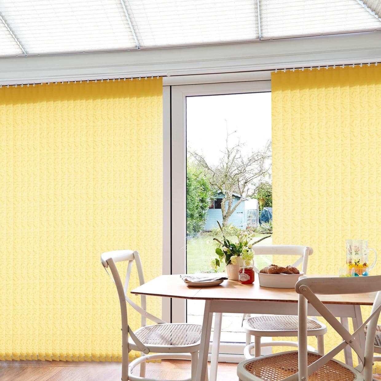 Yellow vertical blinds hung across patio doors, partly drawn to reveal a central glass door; filtering daylight into a bright dining area with table, chairs, and garden view.