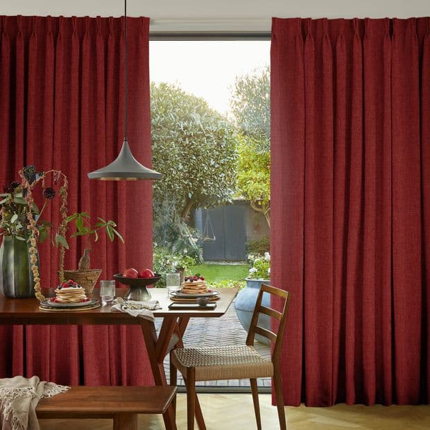 Deep red pleated floor-length curtains, split centrally and textured; partially drawn, framing and filtering daylight; dining room with a table set under a pendant lamp, opening to a green garden.
