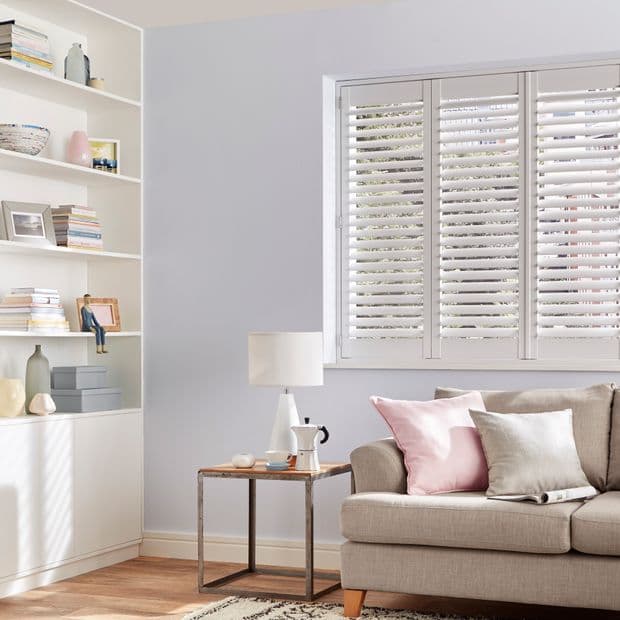 White plantation shutters covering a three-panel window, closed with tilted slats filtering soft daylight; in bright living room beside beige sofa with pink cushions, side table, lamp, and shelves.