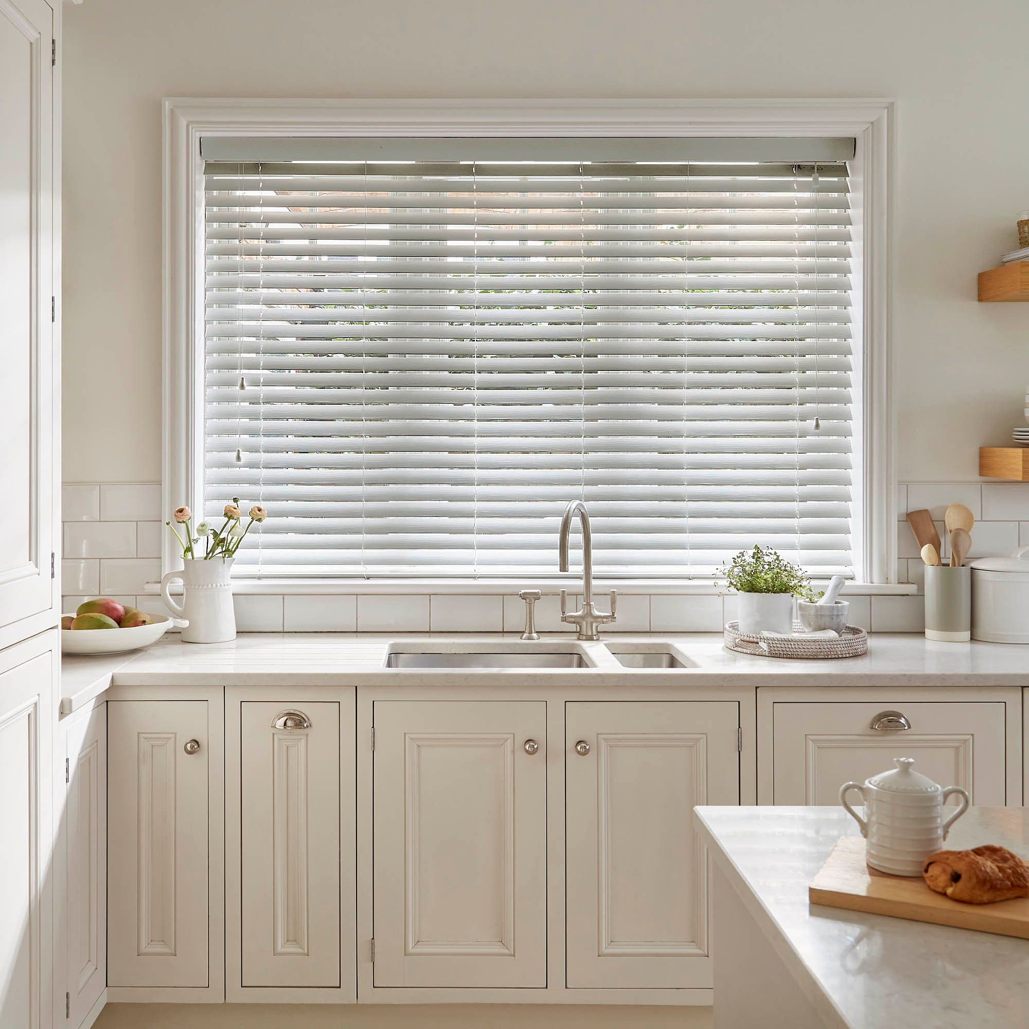 White horizontal slatted blinds across a large window, lowered and slightly tilted to filter soft daylight; set above a farmhouse-style sink in a bright, neutral-toned kitchen.