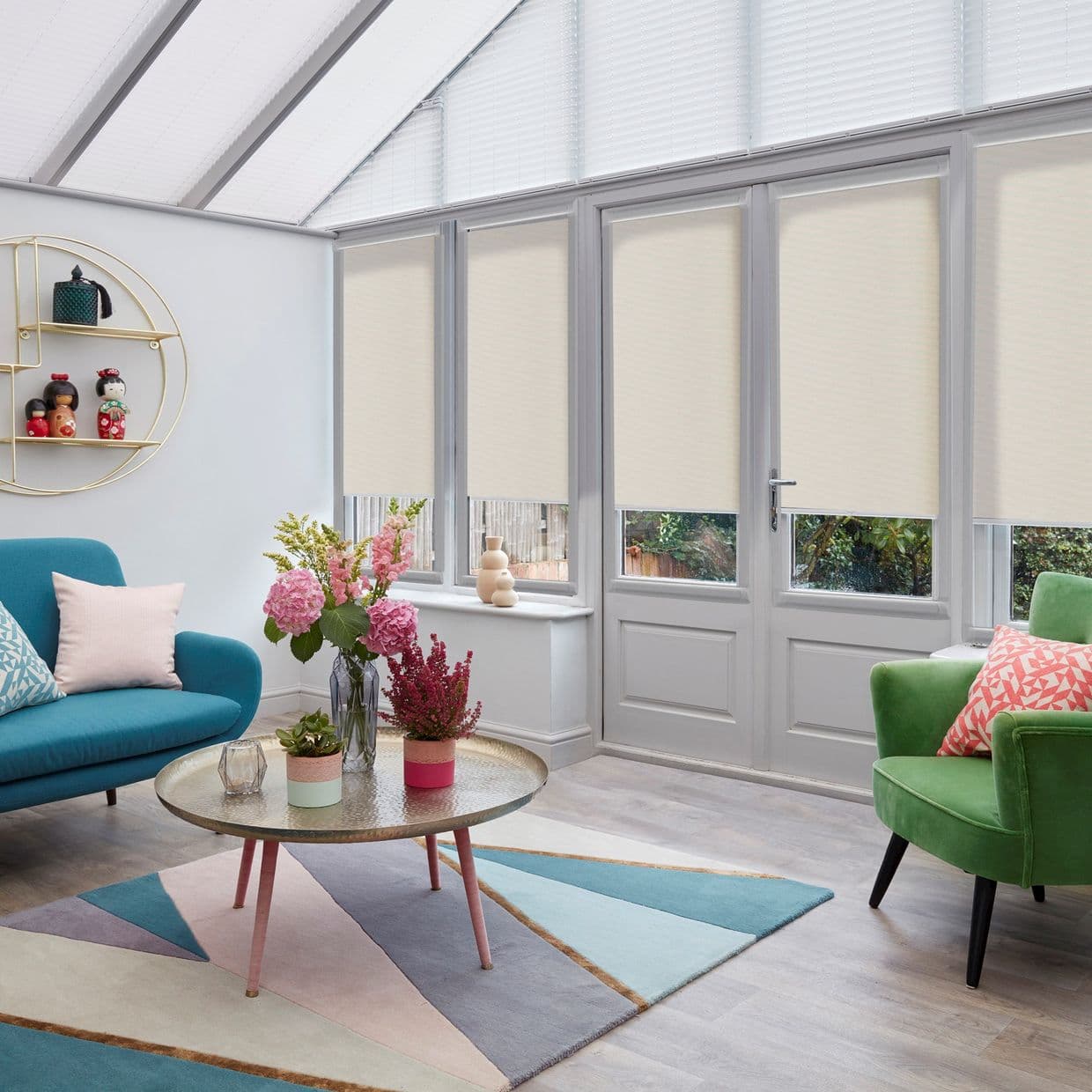Beige roller blinds lowered across tall glazed French doors and windows, lightâ€‘filtering opaque fabric leaving narrow lower gaps; a bright conservatory-style room with colorful sofas, rug and flowers.
