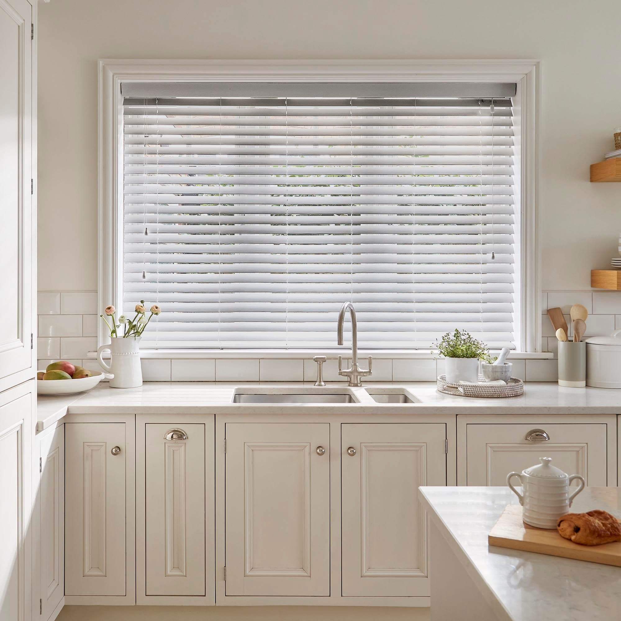 White horizontal Venetian blinds cover a large window, lowered with slats tilted to softly filter daylight; they sit above a farmhouse sink in a bright, neutral kitchen with cream cabinets.