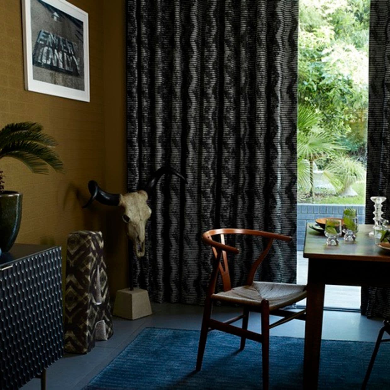 Black patterned floor-to-ceiling curtains cover most of a large sliding door, partially drawn to reveal a sunlit garden; dining area with a wooden chair, set table and a mounted animal skull.