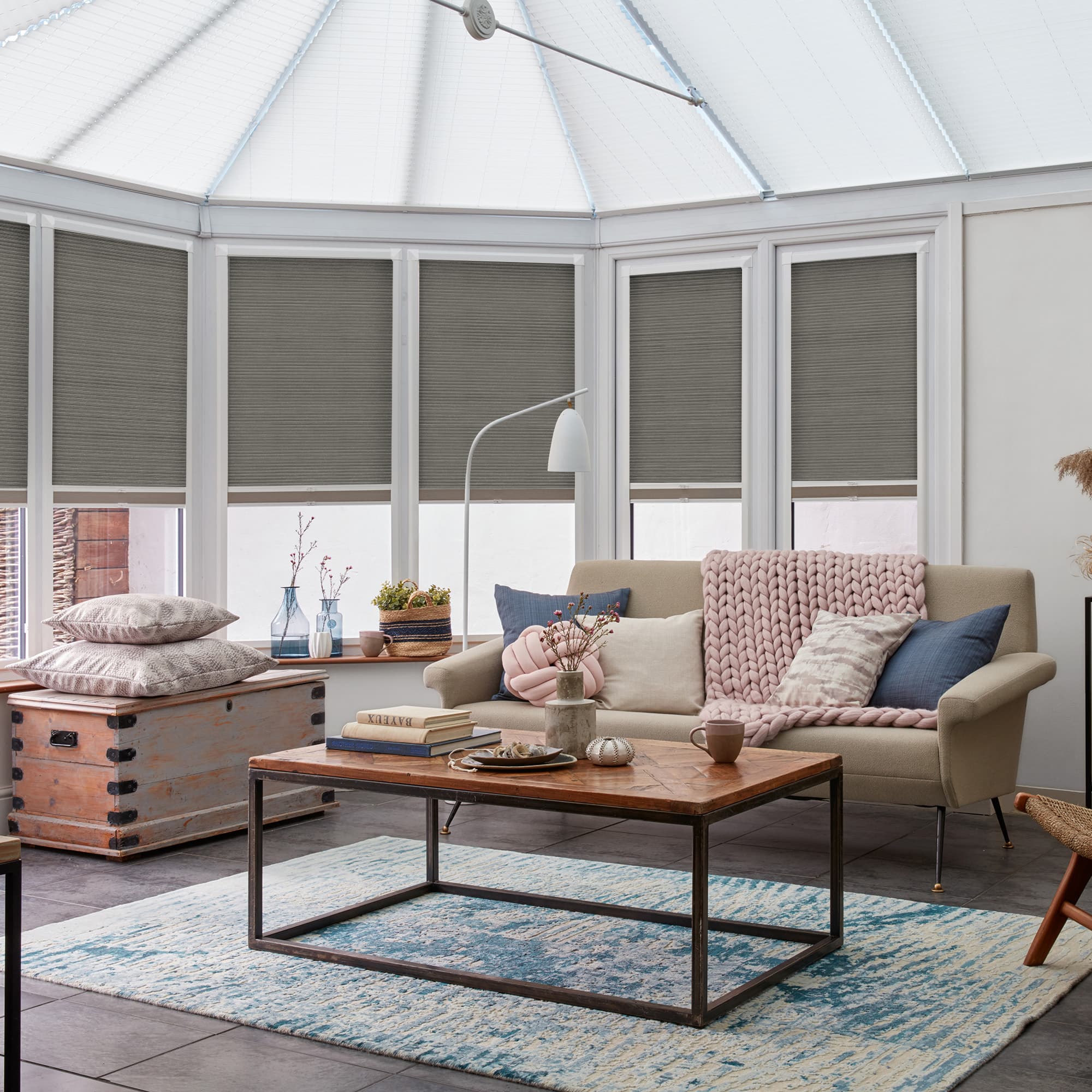 Grey cellular (honeycomb) blinds lowered over tall conservatory windows, softly filtering daylight; cozy sunroom with a beige sofa draped in a chunky pink throw, wooden coffee table and blue rug.
