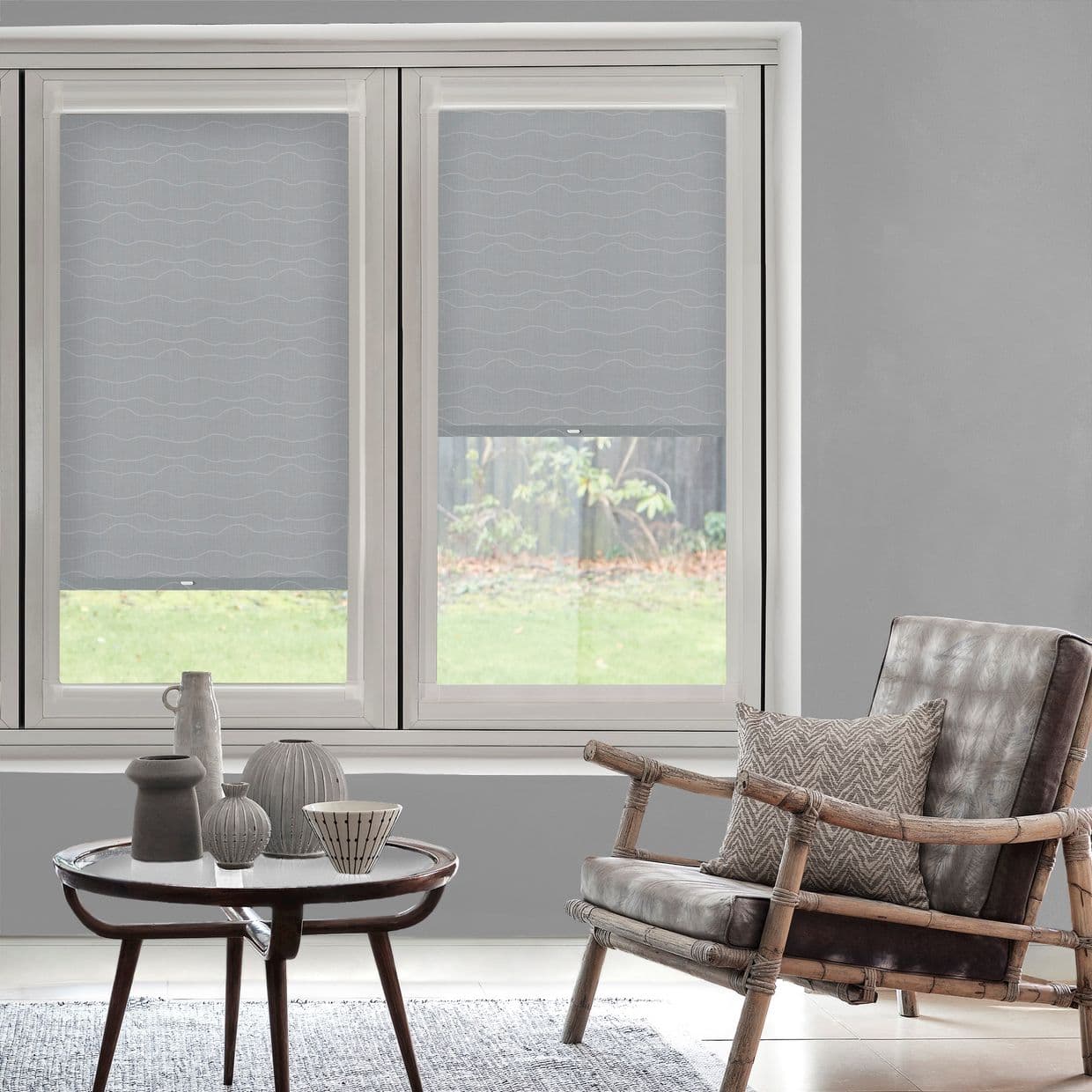 Light-gray patterned roller blinds, lowered partway on twin windows, filtering daylight; neutral-toned living room with rattan-framed leather armchair, patterned cushion and round coffee table with ceramics, grassy yard beyond.