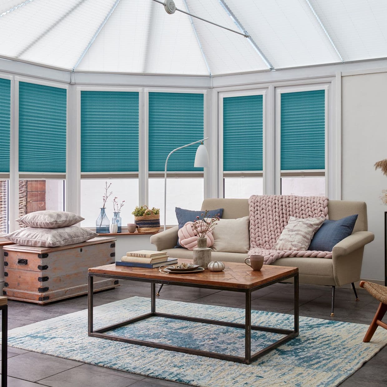 Teal pleated blinds across conservatory-style bay windows, lowered covering upper panes and diffusing daylight; behind a neutral sofa and wooden coffee table in a bright living room.