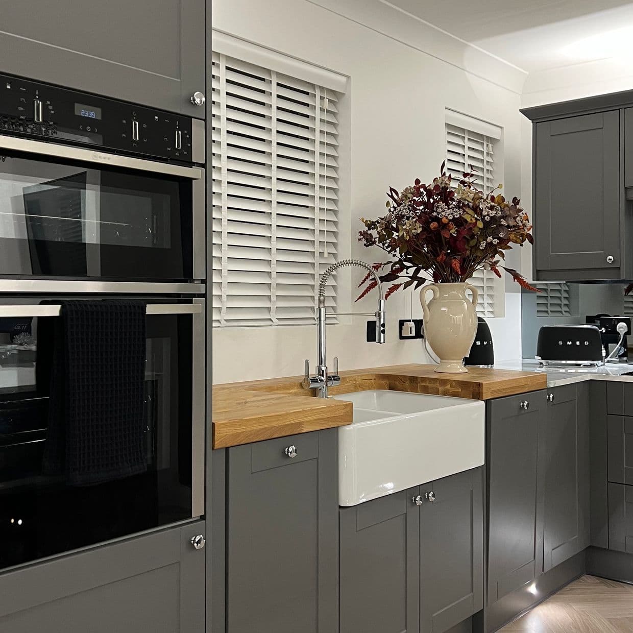 White horizontal slatted blinds over two windows, closed and softly filtering light; foreground farmhouse sink and wooden countertop within grey shaker cabinets, stainless ovens and a vase of autumn flowers.

SMEG
23:33