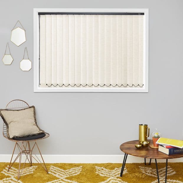 Cream vertical blinds covering a centered window, closed and filtering light with scalloped chain weights at hem; sitting area with grey wall, wire chair, wooden coffee table and mustard rug.