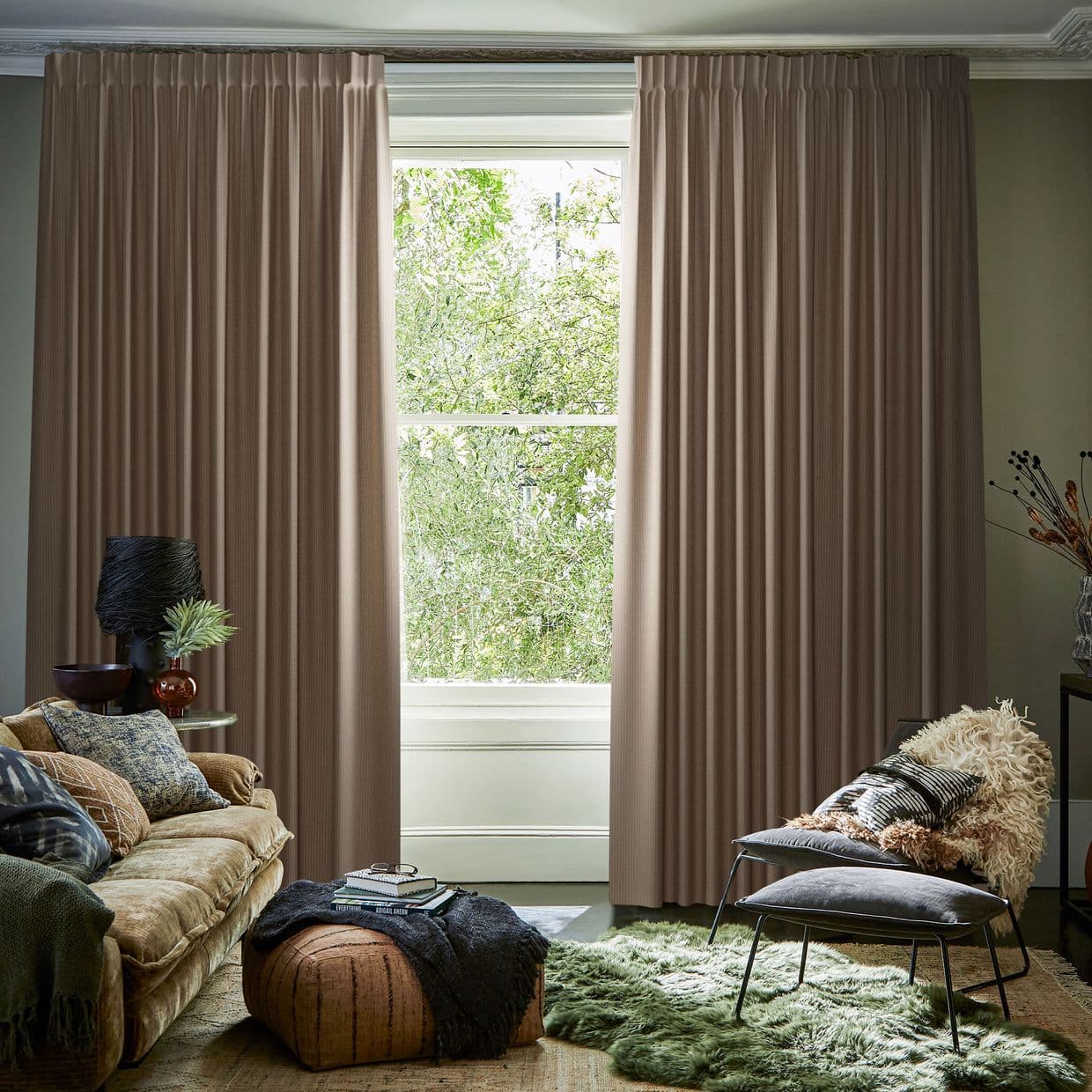 Full-length pleated beige curtains, hung on a ceiling track, mostly closed with a narrow central opening, framing and filtering daylight from a leafy garden visible through a living-room window.