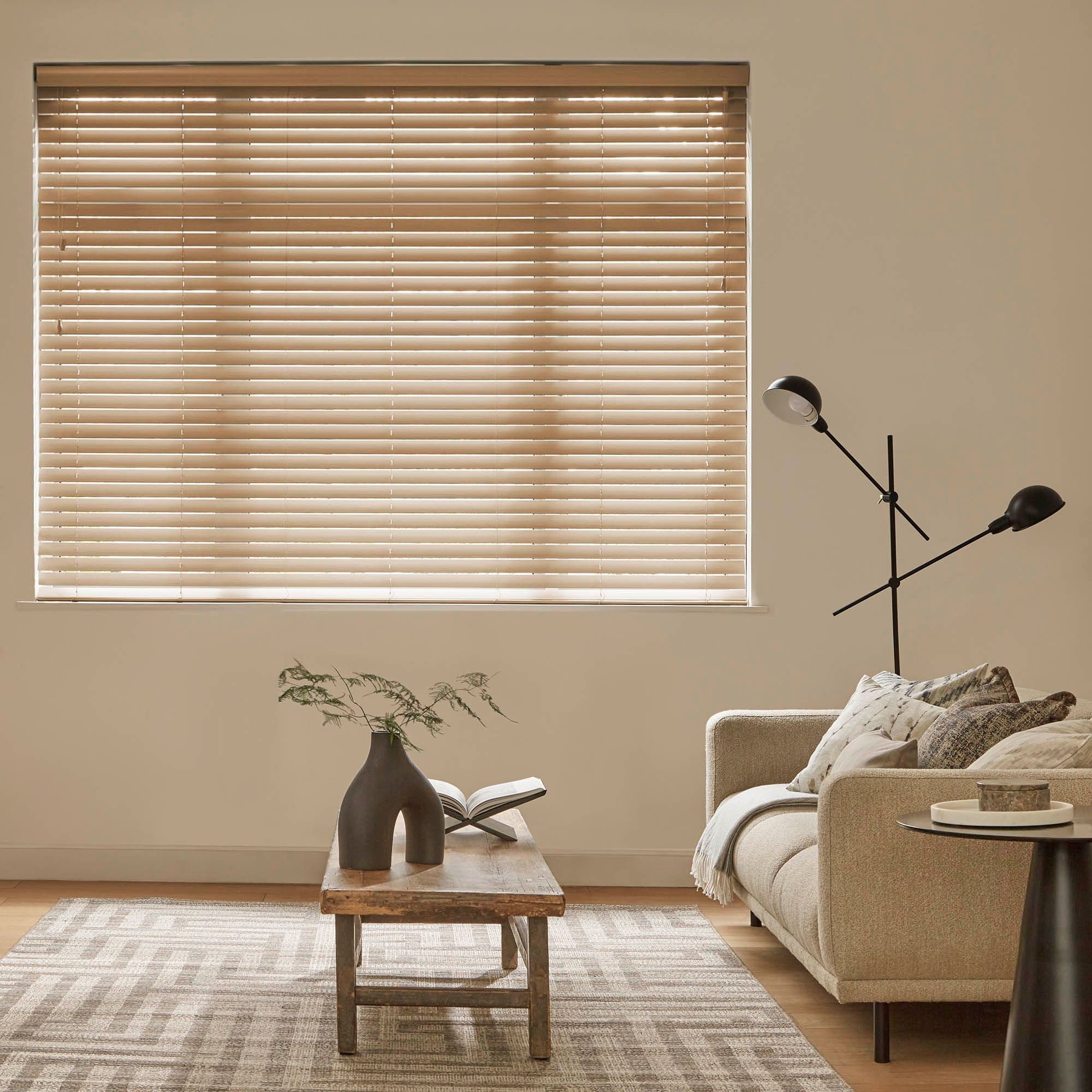Beige horizontal blinds covering a large window, closed and softly filtering daylight; in a neutral-toned living room with a beige sofa, wooden coffee table, patterned rug and modern floor lamp.