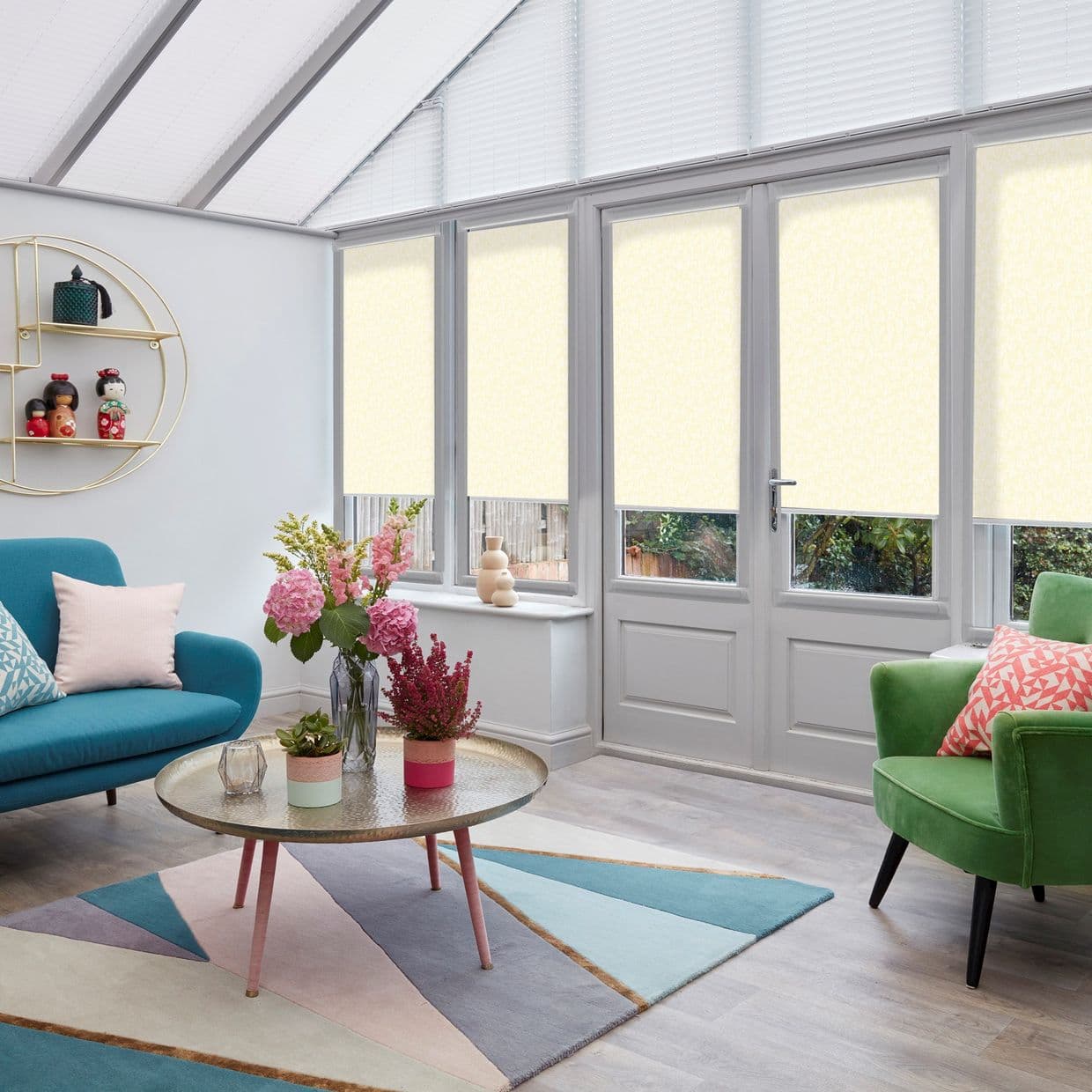 Pale roller blinds over tall windows and a French door, lowered across upper panes to soften daylight; bright conservatory living room with sofa, armchair, coffee table, flowers, and garden view.