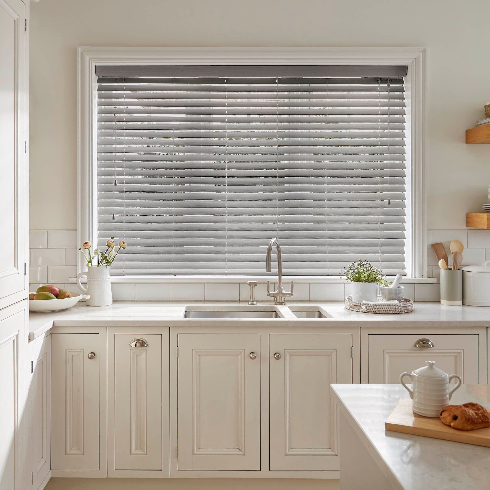 Horizontal Venetian blinds (grey slatted) covering a wide window, closed with slats slightly angled to filter daylight; above a white farmhouse kitchen sink, marble counters, potted herbs and utensils.