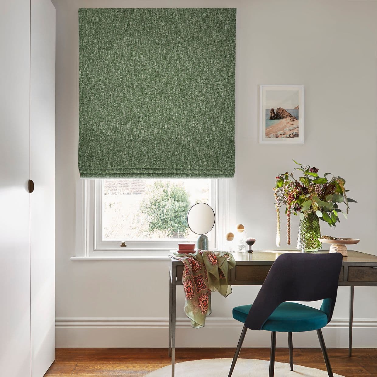 A green textured Roman blind, fully lowered with a slight bottom gap, filtering daylight into a home study; wooden desk, teal chair, round mirror, vase of flowers and framed seaside print.