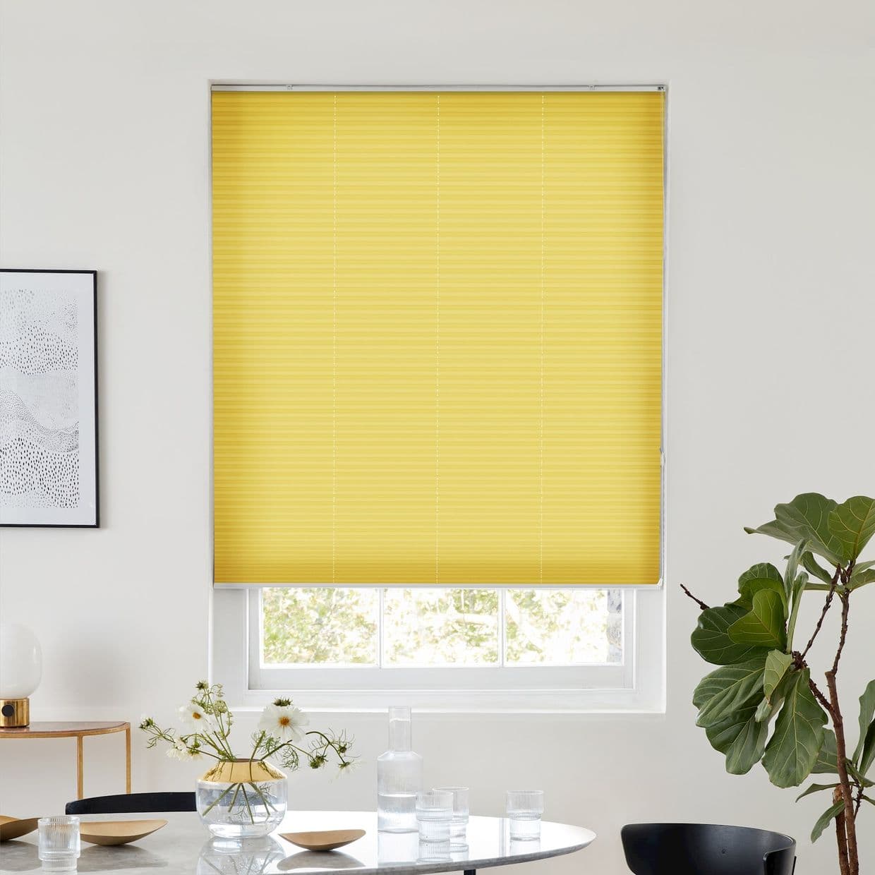 Yellow pleated blind, fully lowered across a window; blocking and softly diffusing daylight in a bright minimalist dining area with round table, vase of flowers, framed art and fiddleâ€‘leaf fig.