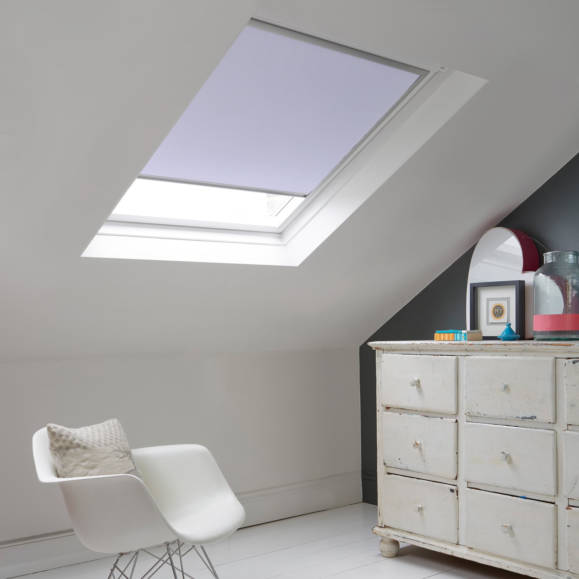 Room with white eaves wall, dark blue-grey wall to right with a shabby chic white drawer unit, a contemporary white chair, white painted floorboards and a skylight blind in pale lilac.