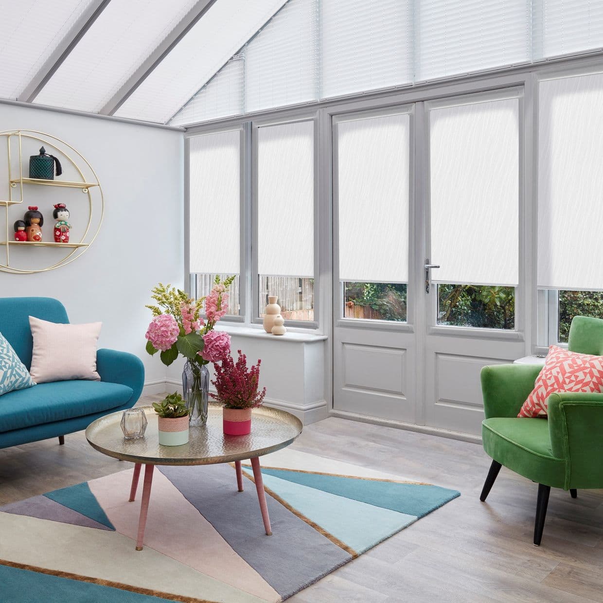 White roller blinds lowered across angled conservatory windows and glazed door, translucently diffusing daylight; sunlit sitting room with teal sofa, green armchair, pastel rug and coffee table with flowers.