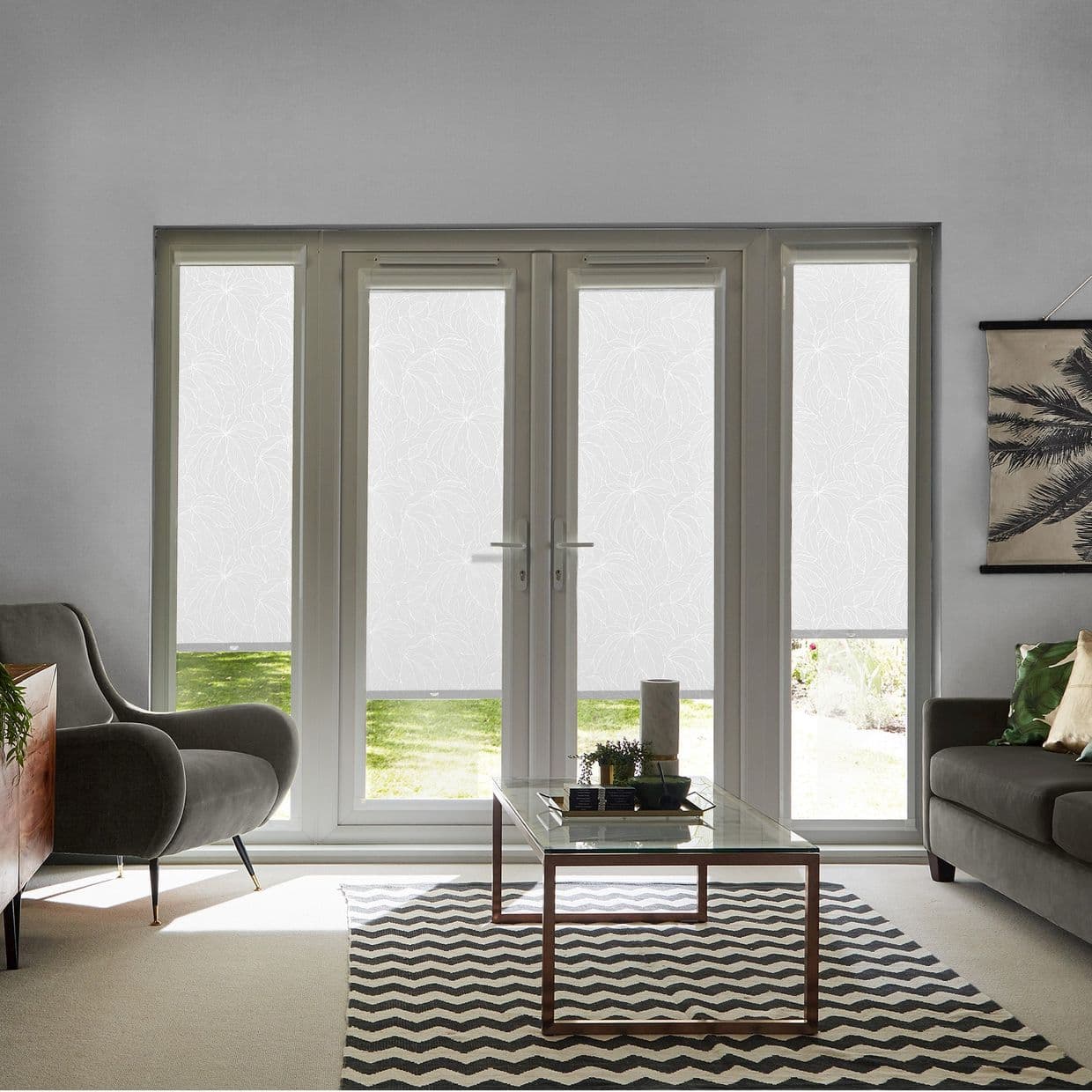 White patterned roller blinds covering French doors and sidelights, lowered to a narrow bottom gap, diffusing daylight and offering privacy; living room with grey sofa, table, rug, garden visible.
