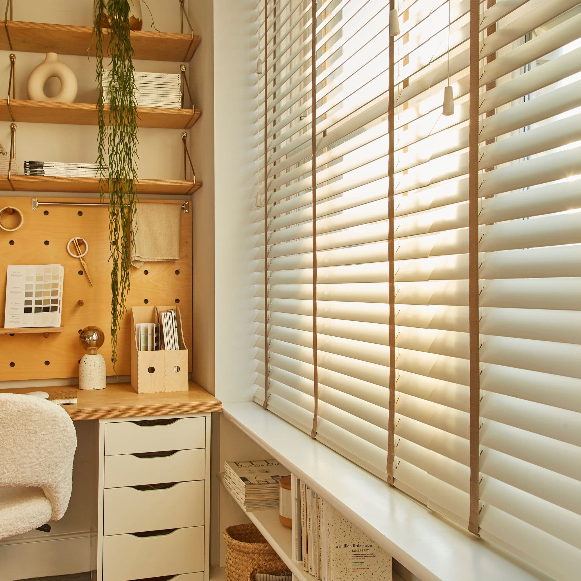 White horizontal venetian blinds covering a window, slats tilted to let warm sunlight filter through; adjacent cozy home office with wooden desk, pegboard, shelves, hanging plant and soft natural lighting.