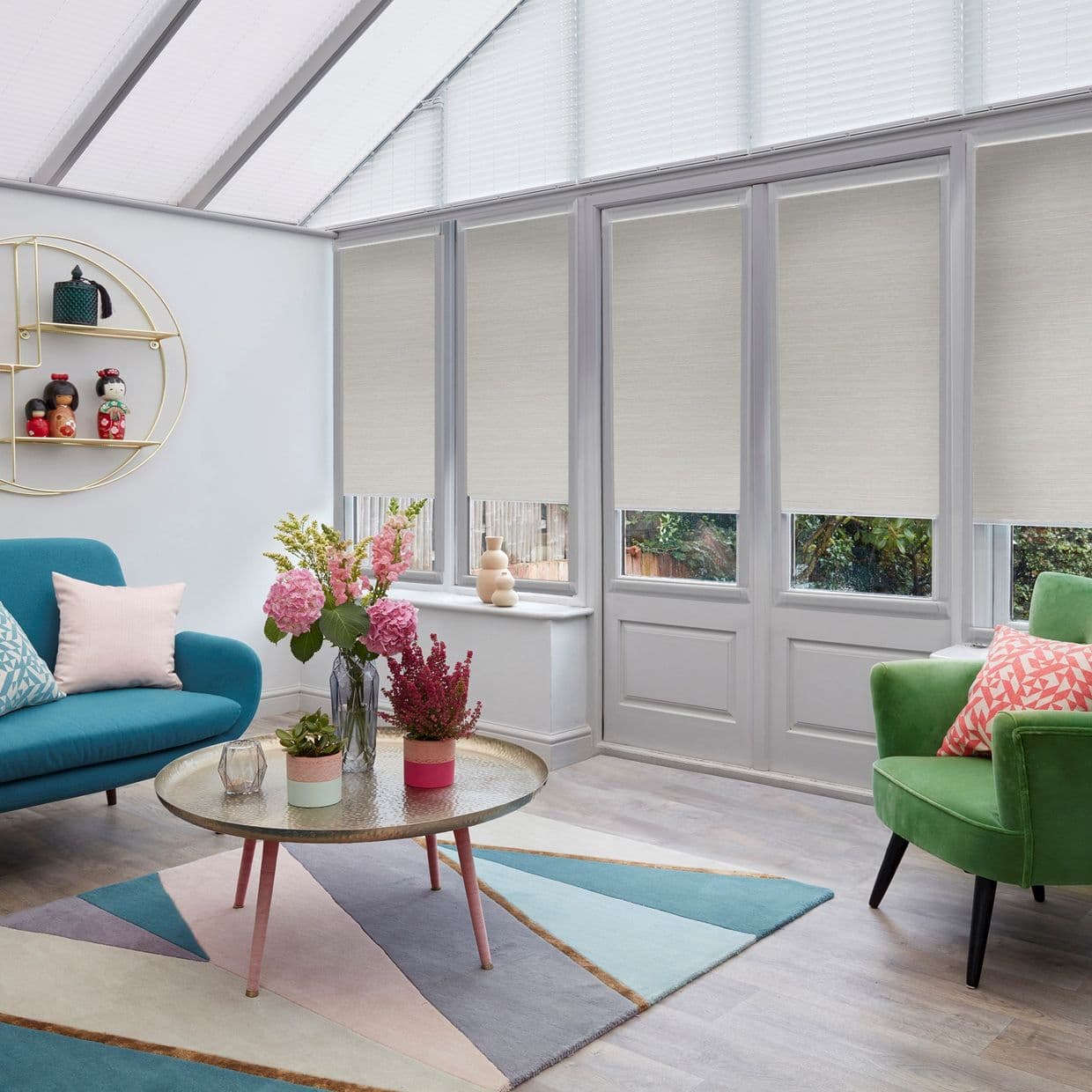 Beige roller blinds, lowered over tall conservatory windows, filtering daylight; contemporary sunroom with teal sofa, green armchair, round coffee table with flowers, pastel geometric rug, soft natural light.