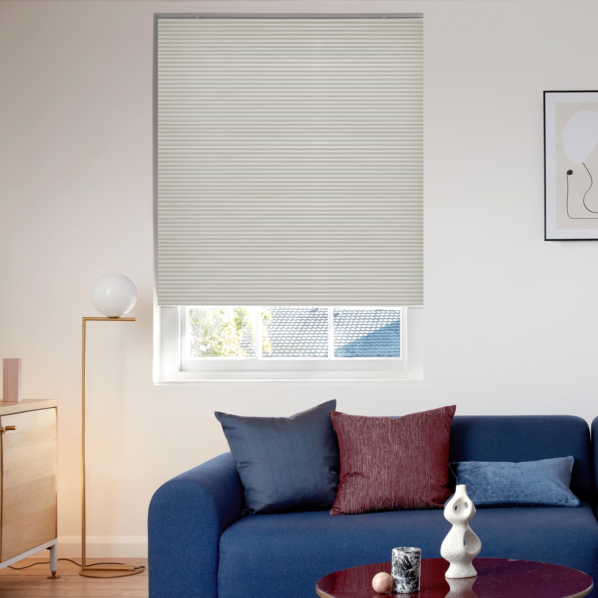 Light grey honeycomb blind lowered covering most of the window, closed with a narrow bottom gap revealing roof and trees; modern living room with blue sofa, cushions, floor lamp, soft daylight.