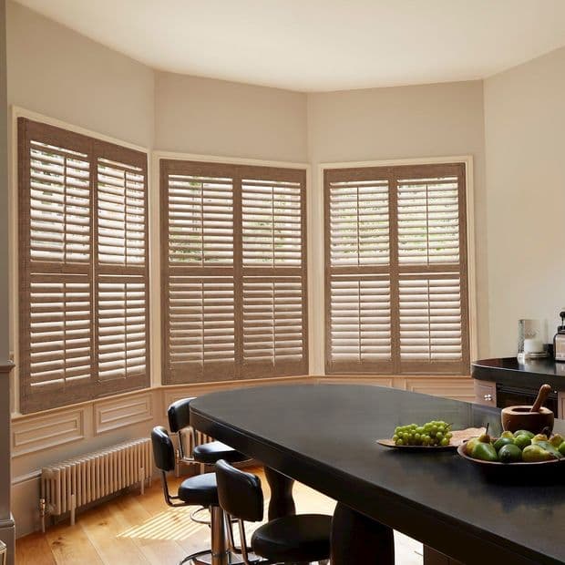 Wooden plantation shutters covering a bay of tall windows, closed with tilted slats filtering soft daylight; overlooking greenery, set above painted wainscoting beside a kitchen island with stools and fruit.