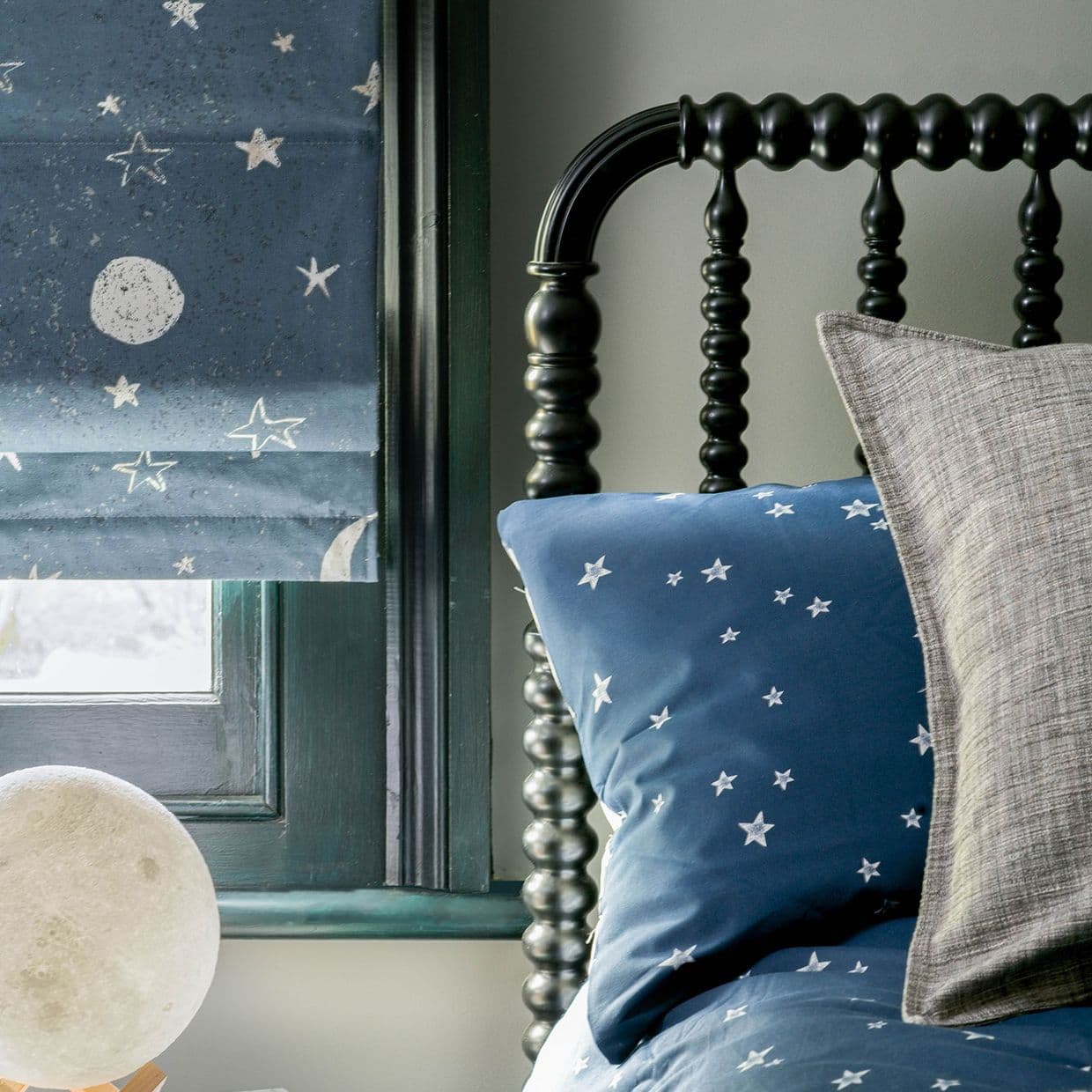 Blue star-patterned Roman shade, partially lowered, printed with moons and stars; filtering soft daylight beside a black turned-spindle headboard with matching star bedding and neutral pillows in a cozy bedroom.