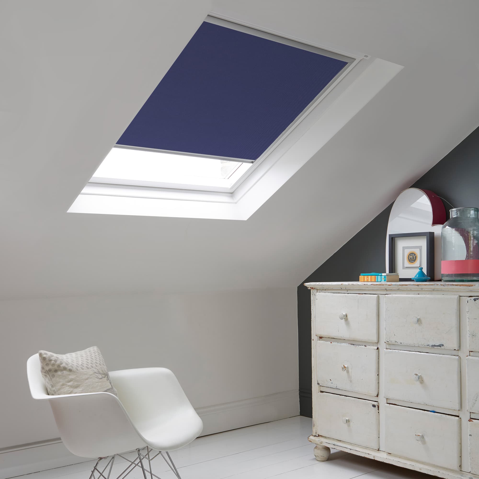 Room with white eaves wall, dark blue-grey wall to right with a shabby chic white drawer unit, a contemporary white chair, white painted floorboards and a skylight blind in ink blue.