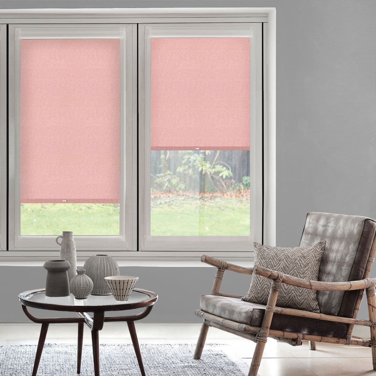 Pink fabric roller blinds on twin windows â€” left lowered, right raised halfway â€” filtering light in living room with rattan armchair, table with ceramics and a grassy yard outside.