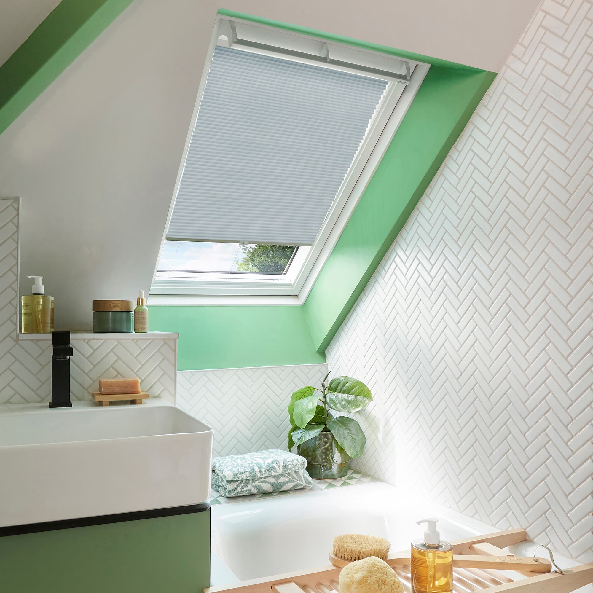 Pleated white cellular blind, lowered over a skylight window, filtering daylight. Bathroom attic space with green-painted trim, white herringbone tiles, sink, tub, potted plant and bath accessories.