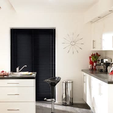 Black horizontal venetian blinds covering a sliding door, fully closed and blocking light â€” in a bright white modern kitchen with stainless stool, trash bin, coffee machine, and red tulips.
