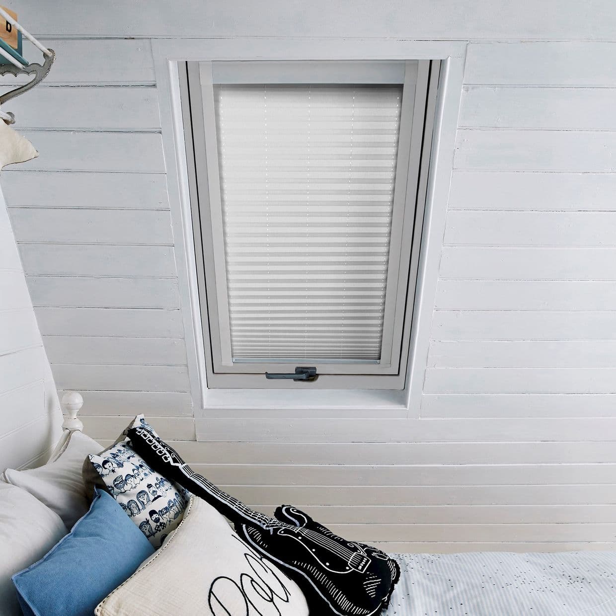 White pleated blinds covering a rectangular skylight, fully closed and softly diffusing daylight; set in a slanted white-painted shiplap ceiling above a bed with pillows and a guitar-shaped cushion.