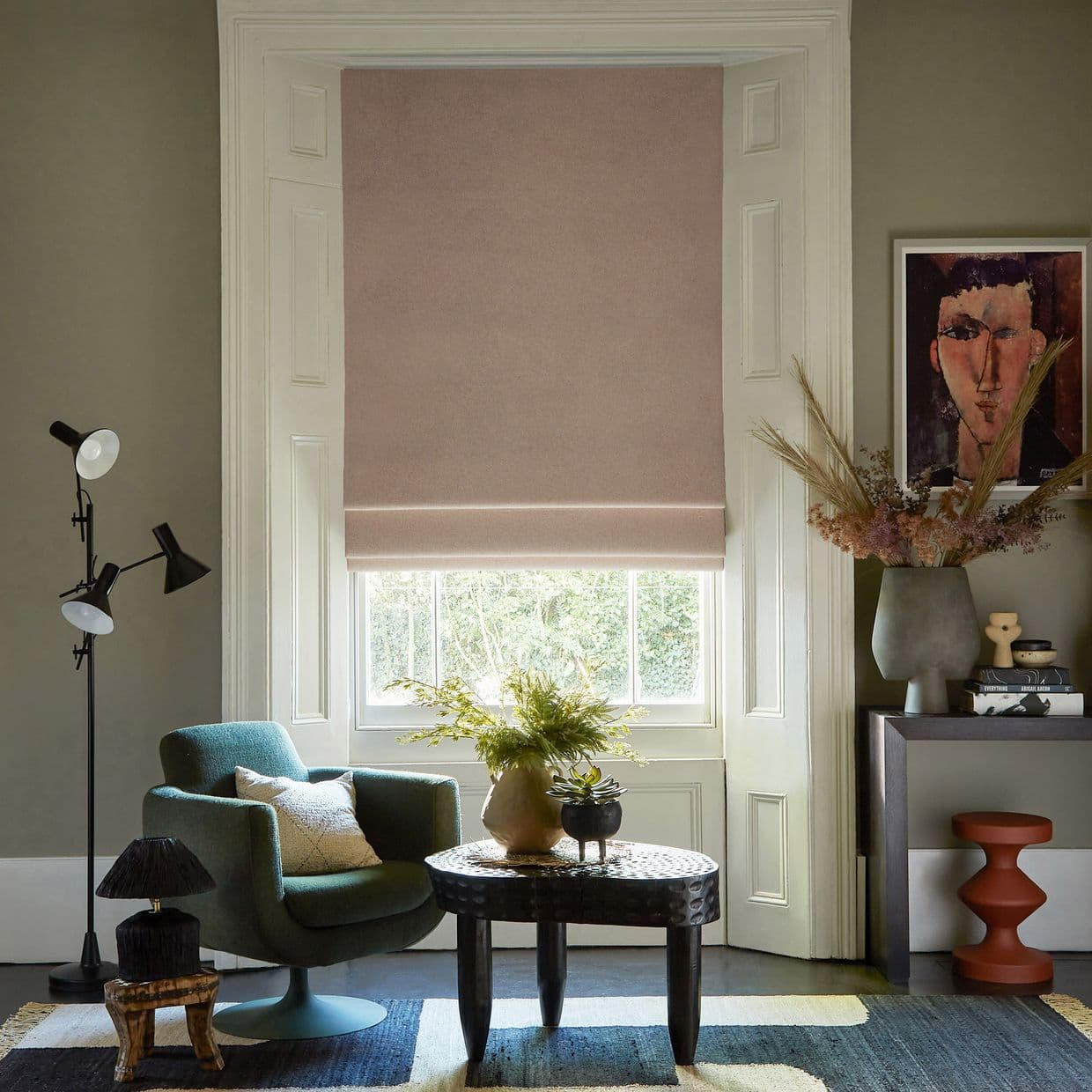 A blush-pink fabric Roman blind, fully lowered with a single fold at the bottom, filtering soft daylight into a cozy sitting room with an armchair, table, plants and framed portrait.