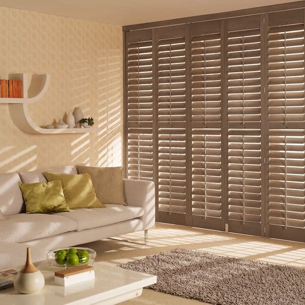 Wooden plantation shutters covering full-height doors, closed with slats angled to filter sunlight; living room with beige sofa, green cushions, cream rug, coffee table, and warm slatted light patterns.