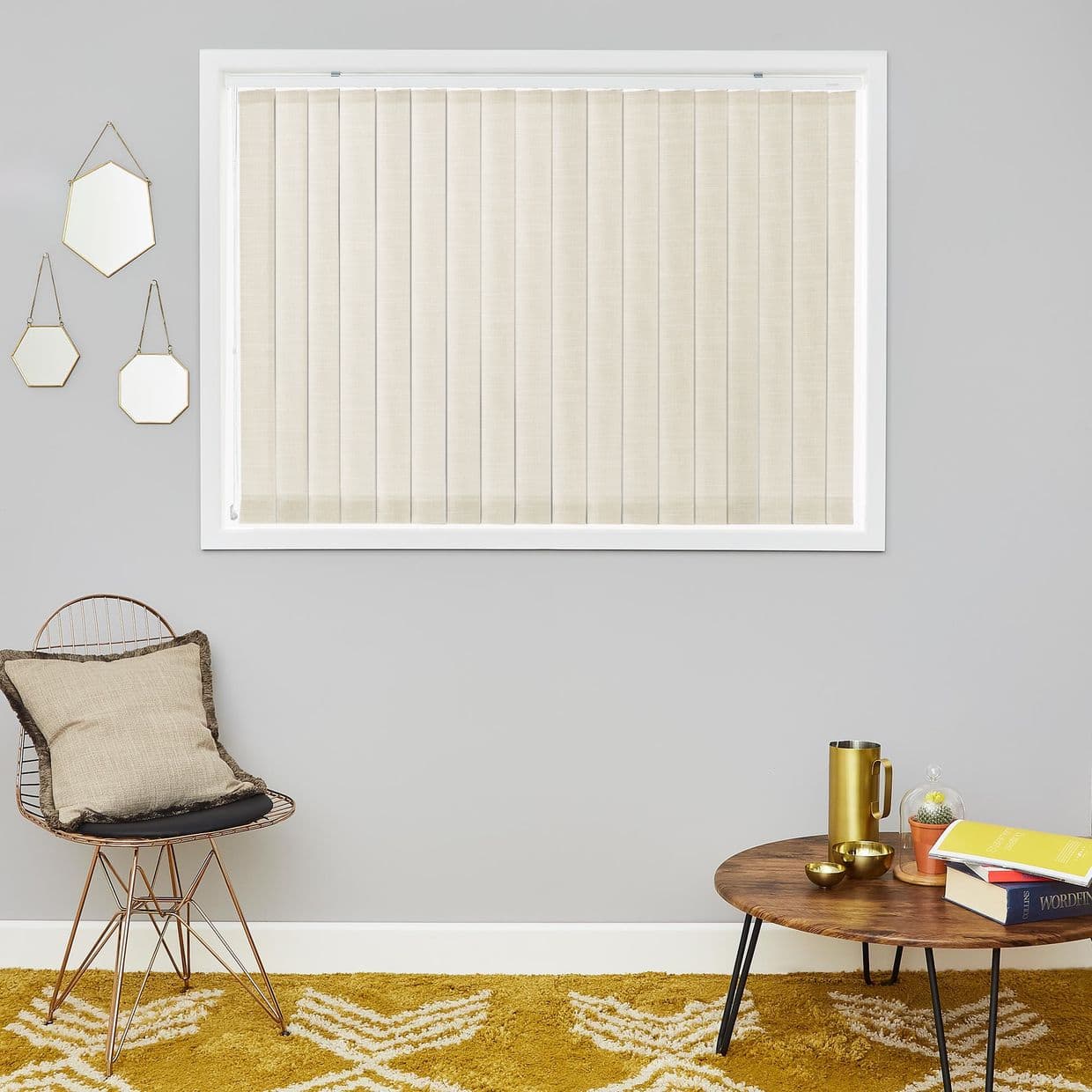 Vertical beige blinds, fully closed and lightly diffusing daylight; set in a white-framed window on a gray wall, above a wire chair with cushion, wooden coffee table and mustard rug.