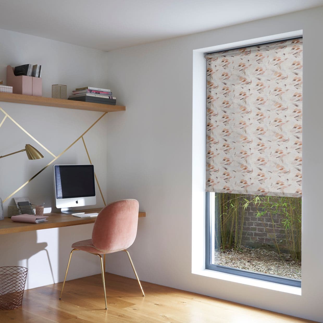 Patterned roller blind with pastel feather print, partially lowered and filtering light, in a modern office beside a wooden desk and pink chair; lower window reveals gravel, bamboo and brick.