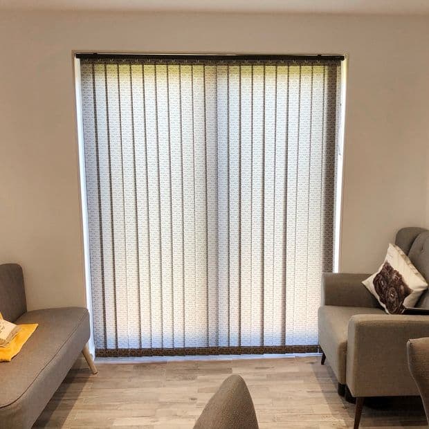 Vertical fabric blinds covering a large sliding window, closed and filtering daylight through patterned slats; modern living room with light wood floor, gray sofas and decorative cushion, soft ambient light.