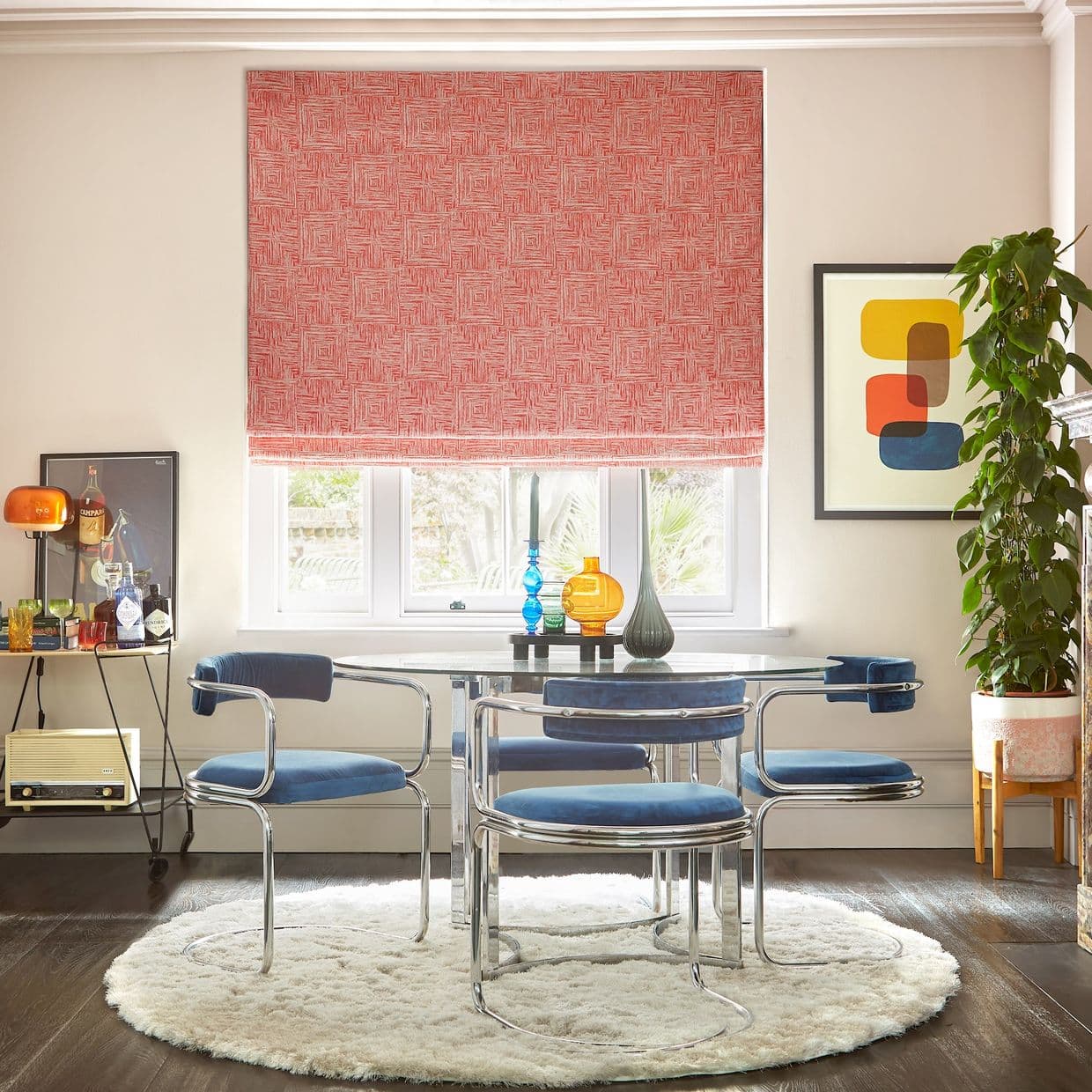 Red patterned Roman blind lowered over a threeâ€‘pane window, filtering daylight; glass dining table with chromeâ€‘framed blue chairs, round shag rug, potted plant and colorful vases; garden view. Campari