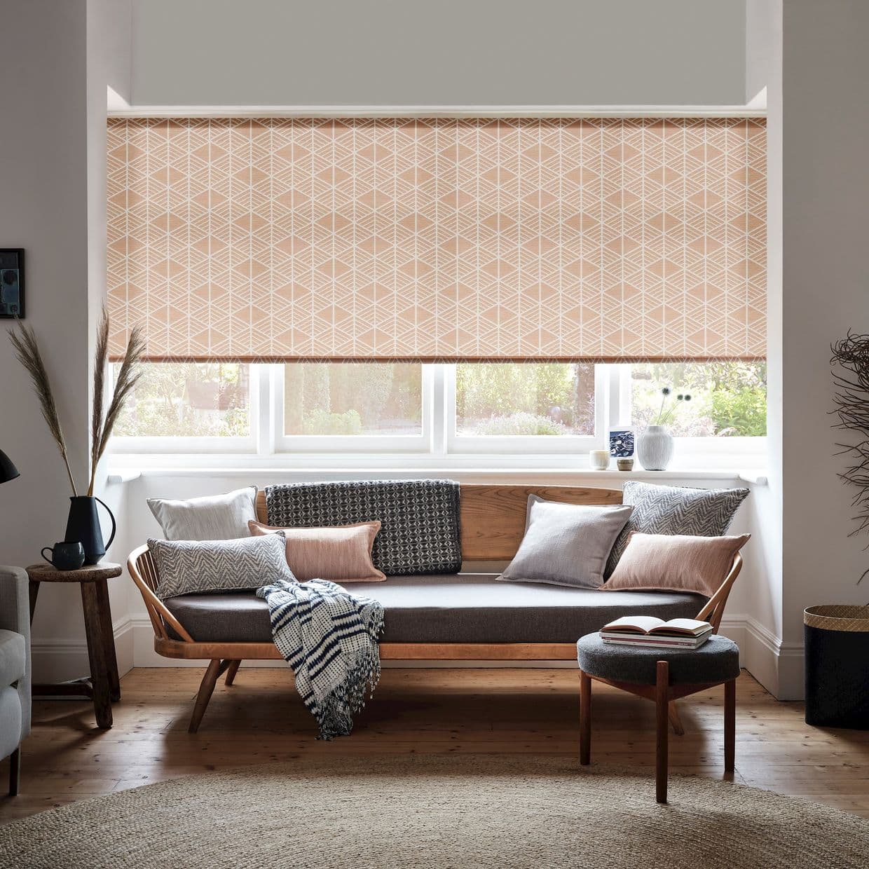 Peach geometric roller blind lowered over a bay window, filtering soft daylight through its lower gap; sits above a wood-framed sofa with cushions in a bright, cozy living room overlooking garden.