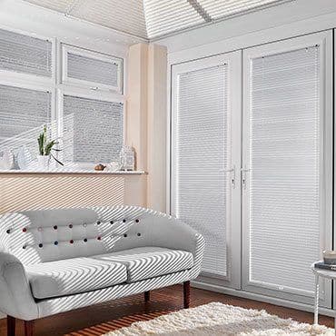 White pleated blinds covering high windows and French doors, closed and filtering sunlight into striped shadows across a curved two-seater sofa and sunlit living room with hardwood floor and rug.