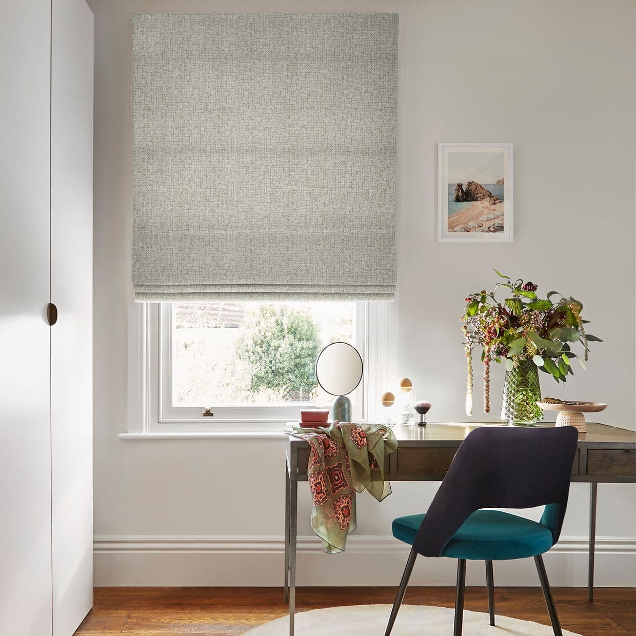 Light-grey patterned Roman blind lowered over window, soft horizontal folds filtering daylight; neutral home office with wooden desk, teal chair, mirror, vase of flowers and framed seaside print.