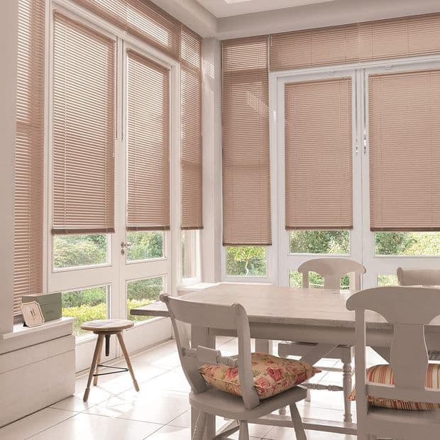 Beige horizontal Venetian blinds, lowered across tall windows and patio doors and filtering soft daylight; wooden dining table and chairs sit in a bright dining area overlooking a green garden.