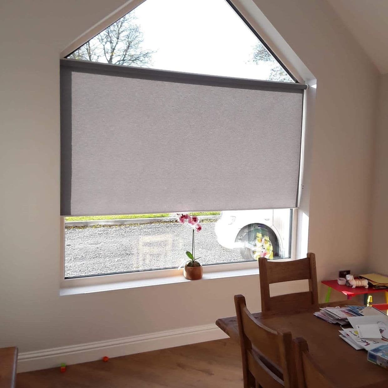 A grey roller blind, lowered to cover most of a tall triangularâ€‘top window, filtering light; dining area with wooden table and chair, small potted orchid on the sill, car visible outside.