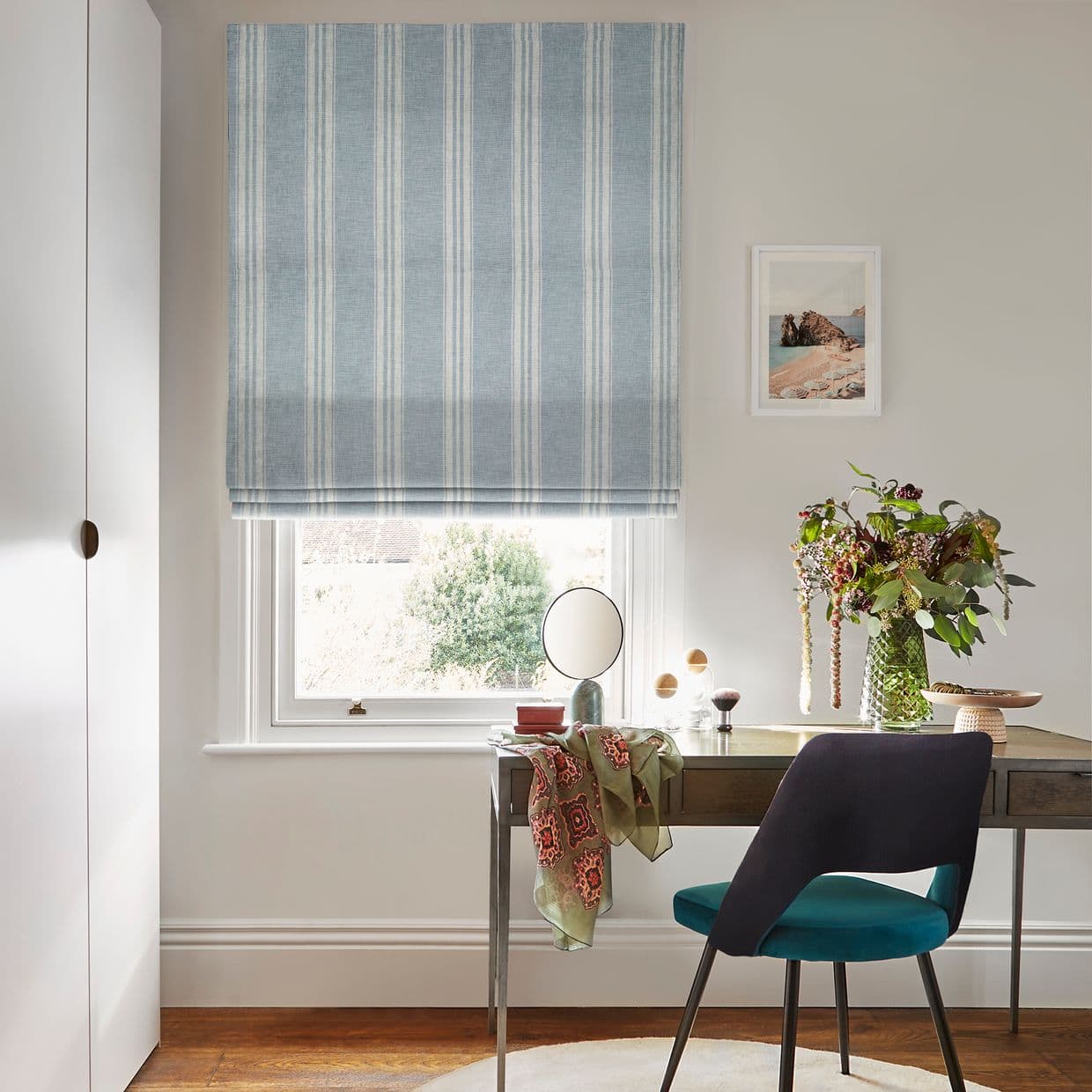 Blue-and-white striped Roman blind, mostly lowered over the window with soft folds, filtering daylight; home office desk beneath with chair, small mirror, floral vase, neutral walls and wooden floor.