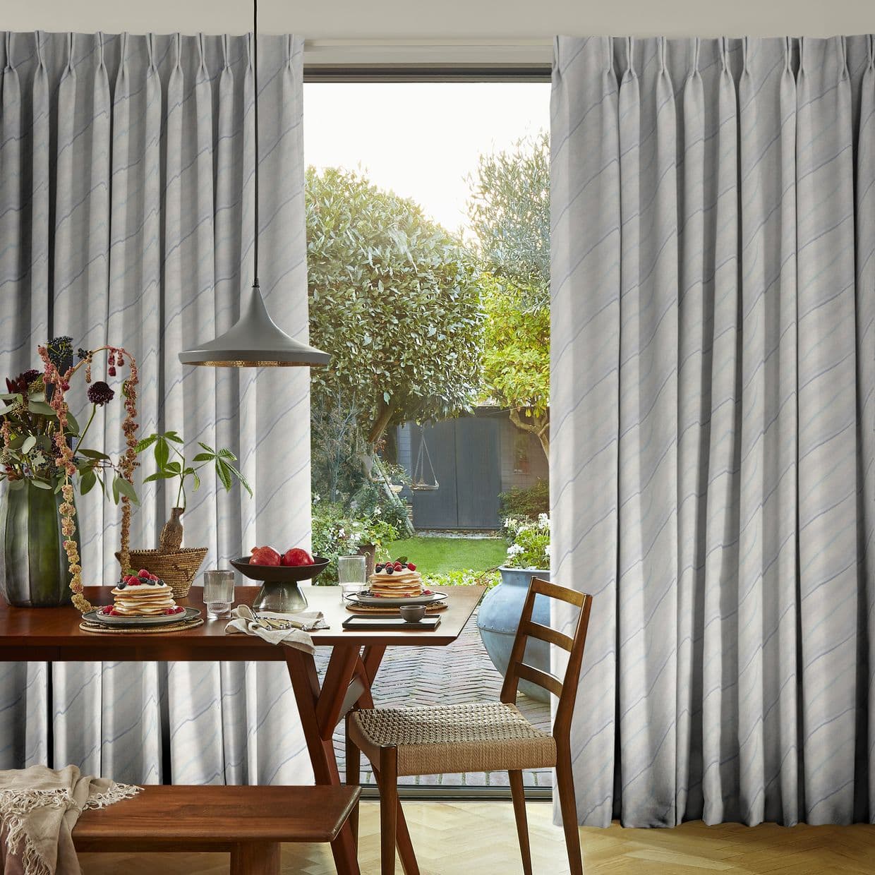 Pleated full-length pale-gray curtains with subtle diagonal stripes, drawn aside to frame the opening; sunlit dining table set for breakfast under a pendant lamp, opening onto a green backyard with trees.