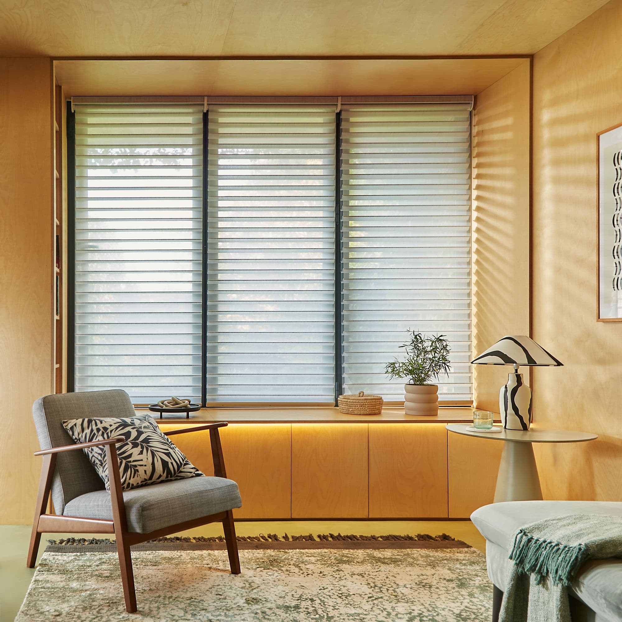 Layered blinds covering a three-panel window, alternating opaque and sheer horizontal bands; filtering soft daylight and casting faint stripes; in a warm wood-paneled living room with armchair and lamp.