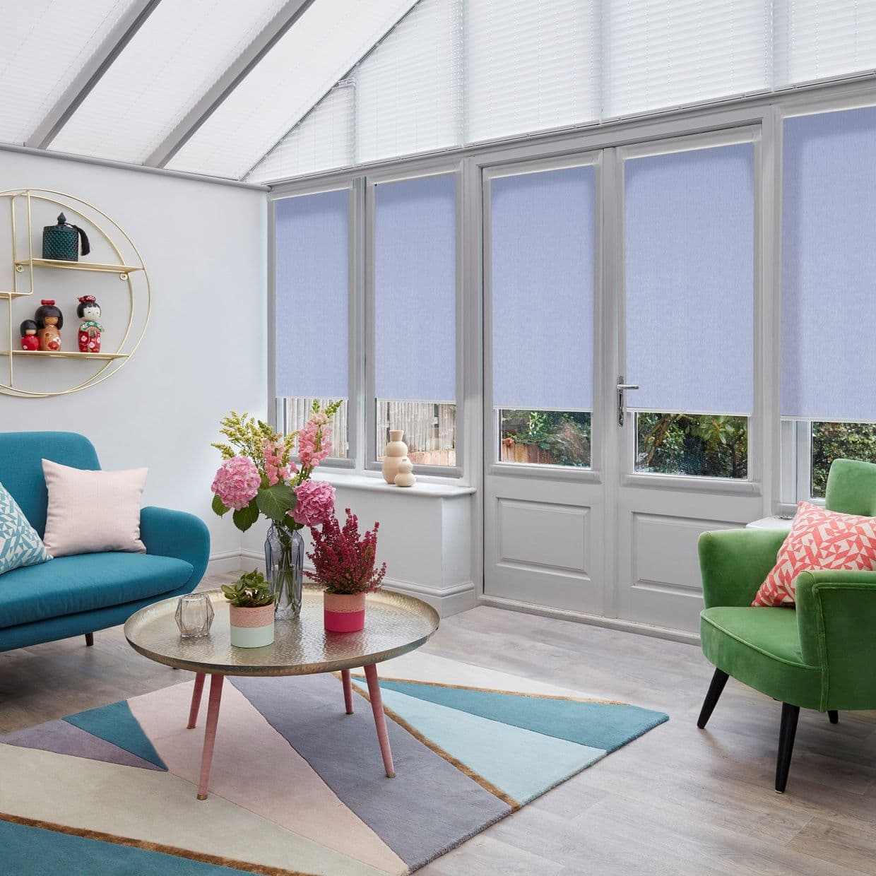 Light-lavender roller blinds covering four tall conservatory windows and door, lowered to mid-height and filtering daylight; modern sunroom with teal sofa, green armchair, round coffee table and floral accents.