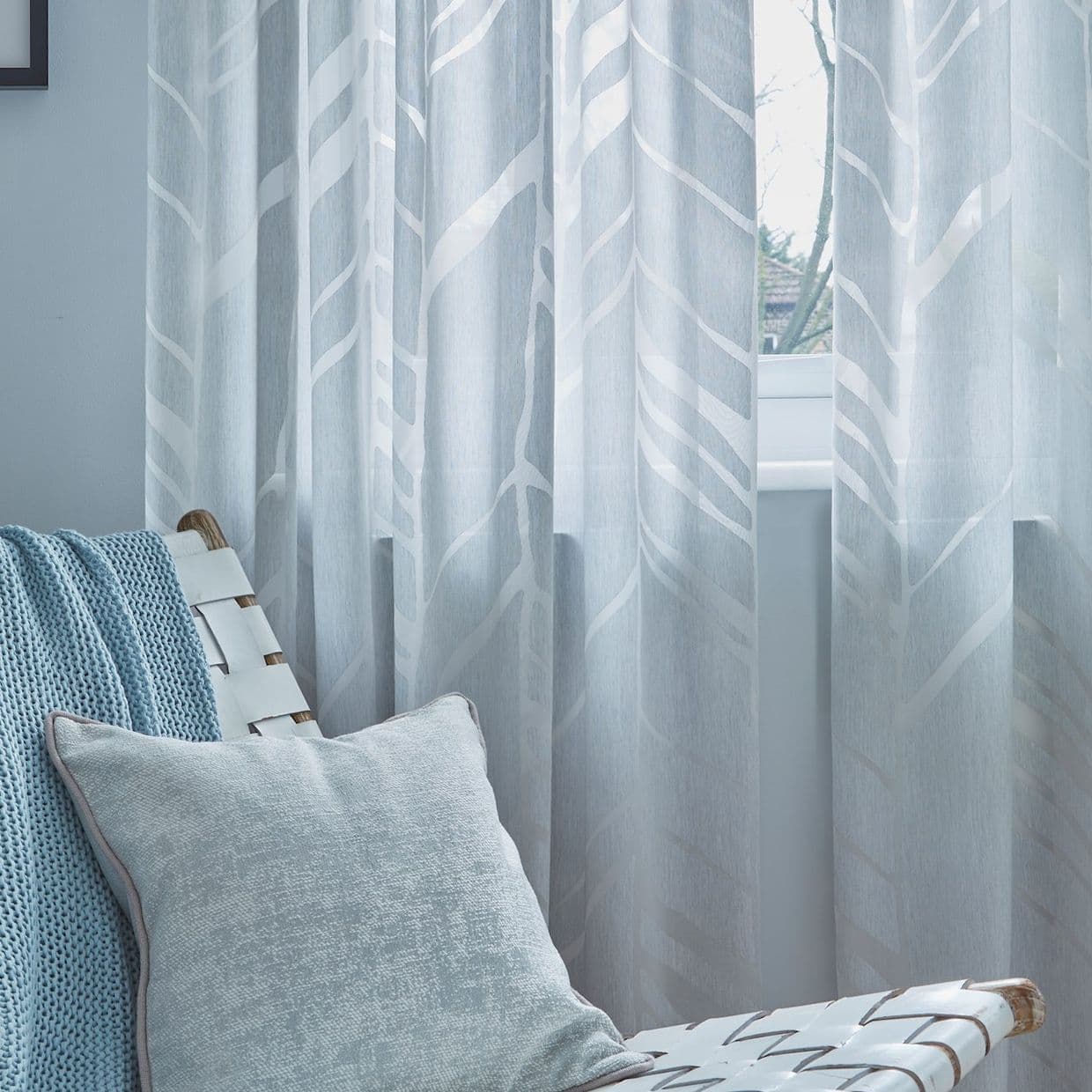 Sheer light-blue curtains with white abstract tree/leaf pattern hang closed over a window, filtering daylight; nearby a woven white chair holds a blue knit throw and pale cushion in a calm, lit room.