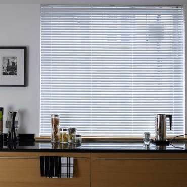 White horizontal Venetian blinds covering a large window, slats mostly closed and filtering diffuse daylight; modern kitchen countertop beneath holds jars, a kettle, utensil holder and wooden cabinets, bright interior.